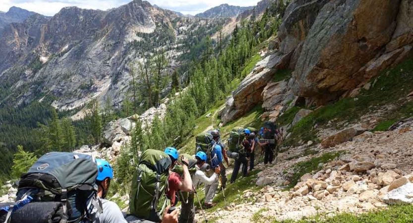 a group of gap year students wearing backpacks and helmets follow a trail up a mountainous landscape